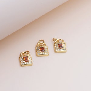 May include: Three gold padlock charms with clear and brown cubic zirconia stones. The charms are on a light pink background.