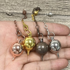 Op de afbeelding: Vier metalen pendels met verschillende kleurafwerkingen: rosé goud, goud, brons en zilver. Elk pendel heeft een ronde bal aan de bovenkant en een puntige punt aan de onderkant.