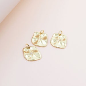 May include: Three gold-colored pendants with an irregular shape and a starburst design in the center. Each pendant has a small loop at the top for attaching to a chain or other jewelry component. The pendants are on a light pink background.