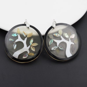 May include: A pair of round earrings with a black background and a white tree design. The tree is decorated with iridescent abalone shell pieces.