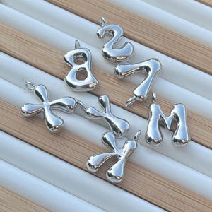 Peut inclure: Un ensemble de six breloques en métal argenté en forme de lettres X, Y, M, 2, 7 et 8. Les breloques sont toutes légèrement déformées et ont une finition brillante.