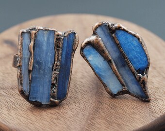 May include: Two rings made of copper and blue kyanite stone. The rings are set on a wooden surface.