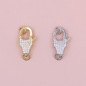 May include: Two lobster claw clasps, one gold-toned and one silver-toned, are displayed against a soft pink background. Both clasps feature a decorative, crystal-studded design on the front, adding a touch of sparkle and elegance to jewelry.