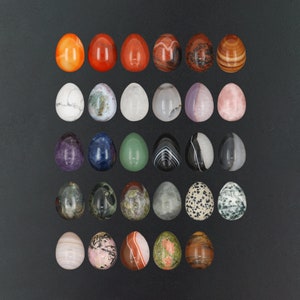 Peut inclure: Une collection de 25 œufs en pierres précieuses polies de différentes couleurs et motifs, notamment rouge, vert, bleu, blanc et noir. Les œufs sont disposés en rangées sur une surface gris foncé.
