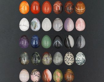 Puede incluir: Una colección de 25 huevos de piedras preciosas pulidas en varios colores y patrones, incluyendo rojo, verde, azul, blanco y negro. Los huevos están dispuestos en filas sobre una superficie gris oscuro.