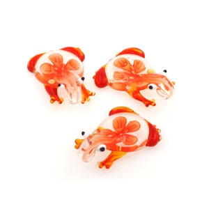 May include: Three glass frog figurines with orange and white floral designs. The frogs are sitting on a white surface.