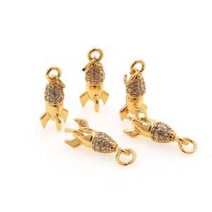 May include: Five gold-colored rocket charms with a sparkling, gem-studded top and a loop for attaching to jewelry. The rocket charms have three fins and a circular loop at the top. The charms are on a white background.