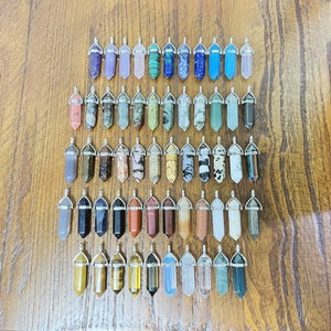May include: A collection of 50 crystal point pendants with silver caps. The pendants are arranged in rows on a wooden surface. The crystals are various colors and patterns, including clear, white, black, brown, green, blue, and purple.
