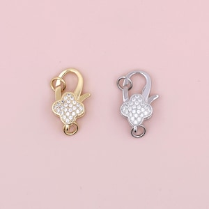 Könnte beinhalten: Zwei Verschlüsse in Silber- und Goldtönen mit einem Blumendesign. Die Blume ist mit klaren Strasssteinen besetzt.