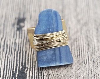 May include: A handcrafted ring featuring a rectangular, light blue stone wrapped with gold-colored wire. The ring has a wide, gold-colored band. The stone's shape is irregular, adding to its unique, artistic appeal. The ring is set against a neutral, textured background.