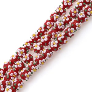 May include: A strand of red glass beads with white flower designs and green accents. The beads are oval shaped and have a glossy finish.
