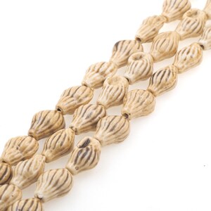 May include: A strand of ceramic beads in a light brown color with a textured, ribbed surface. The beads are shaped like teardrops and are strung together on a clear thread.