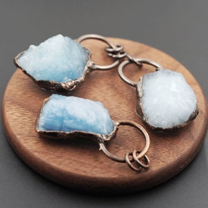 May include: Three blue aquamarine crystal pendants with copper wire wrapped around them. The pendants are on a wooden surface.