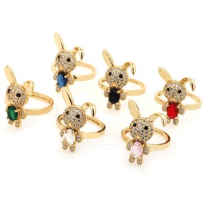 May include: Six gold-tone rings featuring a rabbit design with a different colored gemstone in the center of each rabbit. The gemstones are blue, black, green, clear, pink, and red.