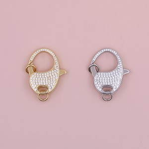 Könnte beinhalten: Zwei silberne und goldene Hummerverschlüsse mit klaren Zirkonia-Steinen. Die Verschlüsse sind beide oval geformt und haben eine kleine Schlaufe an der Seite.