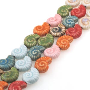 Könnte beinhalten: Eine Reihe bunter Keramikperlen in Form von Muscheln. Die Perlen sind in verschiedenen Blautönen, Grüntönen, Rosa, Orange und Rot.