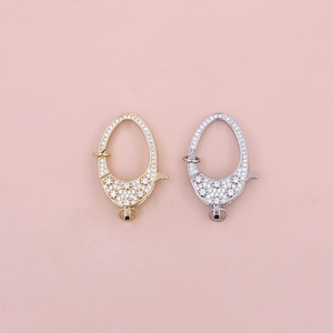 Könnte beinhalten: Zwei ovale Verschlüsse mit einem klaren Kristalldesign. Ein Verschluss ist goldfarben und der andere silberfarben.