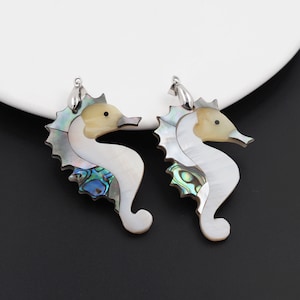 Könnte beinhalten: Ein Paar silberfarbene Ohrringe mit einem Seepferdchen-Design aus irisierendem Abalone-Schalenmaterial. Die Seepferdchen sind weiß mit einem gelben Gesicht und blauen und grünen Akzenten.