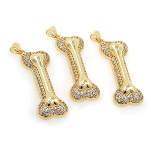 May include: Three gold-tone bone-shaped pendants with clear cubic zirconia stones. The pendants are on a white background.