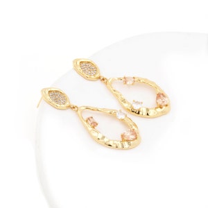 May include: Gold tone dangle earrings with a textured oval shape and clear and peach coloured stones.