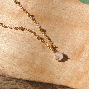 May include: A delicate gold chain necklace with a small, light pink gemstone pendant. The necklace is displayed on a wooden surface.