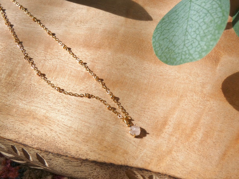 May include: A delicate gold chain necklace with a small, light pink gemstone pendant. The necklace is displayed on a wooden surface.