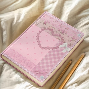 May include: A pink journal with a heart-shaped cutout and floral patterns. The cover features a polka dot design, lace trim, and a gingham pattern. Two pencils rest beside the journal. The journal has the text "zara eclectica".