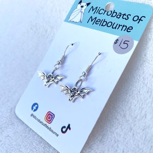 May include: Silver bat earrings on a white card with "Microbats of Melbourne" printed on it. The earrings feature a detailed bat design. Social media icons for Facebook, Instagram, and TikTok are also visible on the card.