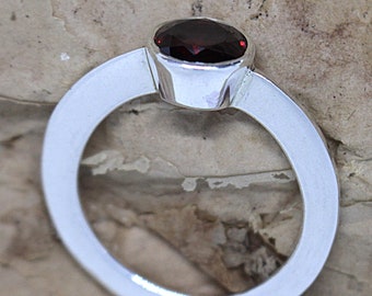 Pode incluir: Um anel de prata com uma gema oval de cor vermelha escura engastada em uma moldura de bisel.