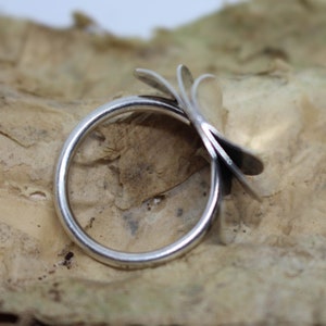 K&ouml;nnte beinhalten: Ein silberner Ring mit einem einzigartigen Design aus mehreren &uuml;berlappenden Bl&uuml;tenbl&auml;ttern, die einer Blume &auml;hneln. Der Ring wird auf einer strukturierten braunen Oberfl&auml;che gezeigt.