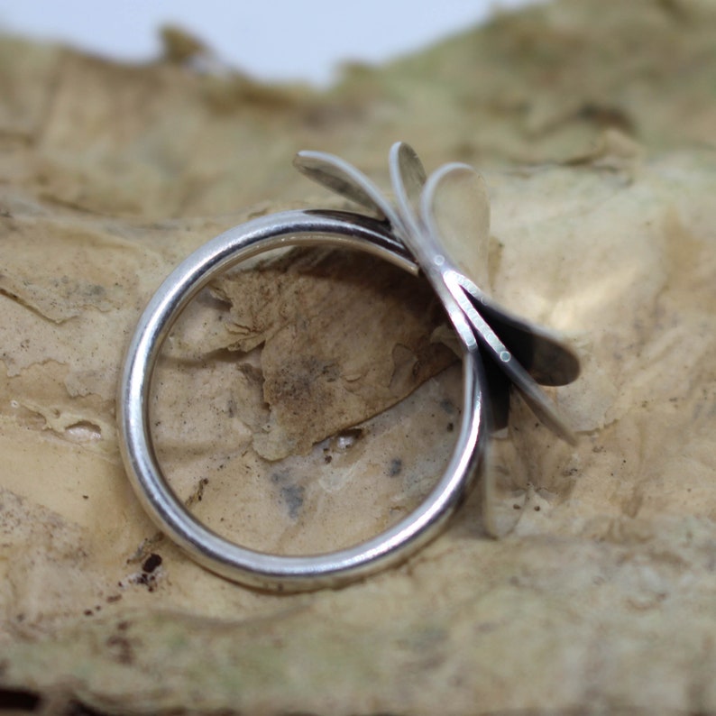 K&ouml;nnte beinhalten: Ein silberner Ring mit einem einzigartigen Design aus mehreren &uuml;berlappenden Bl&uuml;tenbl&auml;ttern, die einer Blume &auml;hneln. Der Ring wird auf einer strukturierten braunen Oberfl&auml;che gezeigt.