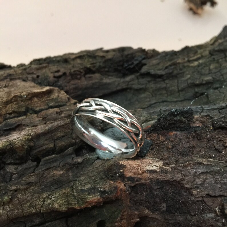 May include: A silver ring with a braided design. The ring is on a piece of wood.