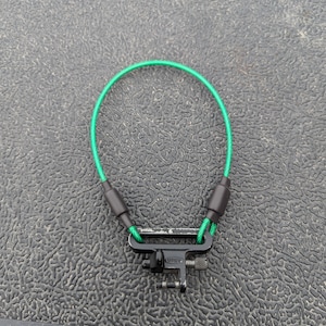 May include: A green and black sling with a metal swivel clip. The sling is made of a braided cord and has a black plastic coating on the ends.