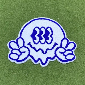 May include: White felt patch with a blue outline of a smiling face with three eyes and peace signs on each side.