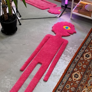 Könnte beinhalten: Ein leuchtend pinkfarbener Teppich in Form einer stilisierten Figur mit langen Gliedmaßen und einem runden Kopf. Der Teppich hat ein großes gelbes und blaues Auge. Der Teppich liegt auf einem grauen Betonboden, mit einem gemusterten Teppich und einem Spiegel im Hintergrund.