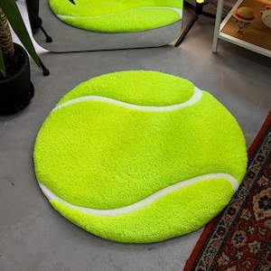 May include: A round, bright green tennis ball rug with white curved stripes. The rug is made of a soft, plush material and is placed on a gray floor. The rug is a fun and playful addition to any room.