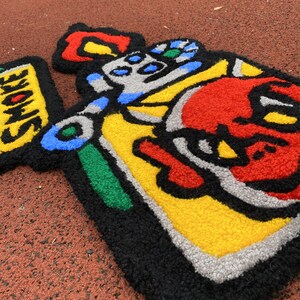 K&ouml;nnte beinhalten: Ein farbenfroher, handgefertigter Teppich mit einem Cartoon-Design. Der Teppich hat einen schwarzen Umriss mit roten, gelben, blauen, gr&uuml;nen und grauen Abschnitten. Das Wort "SMOKE" ist auf gelbem Hintergrund zu sehen. Der Teppich liegt auf einer roten Oberfl&auml;che.