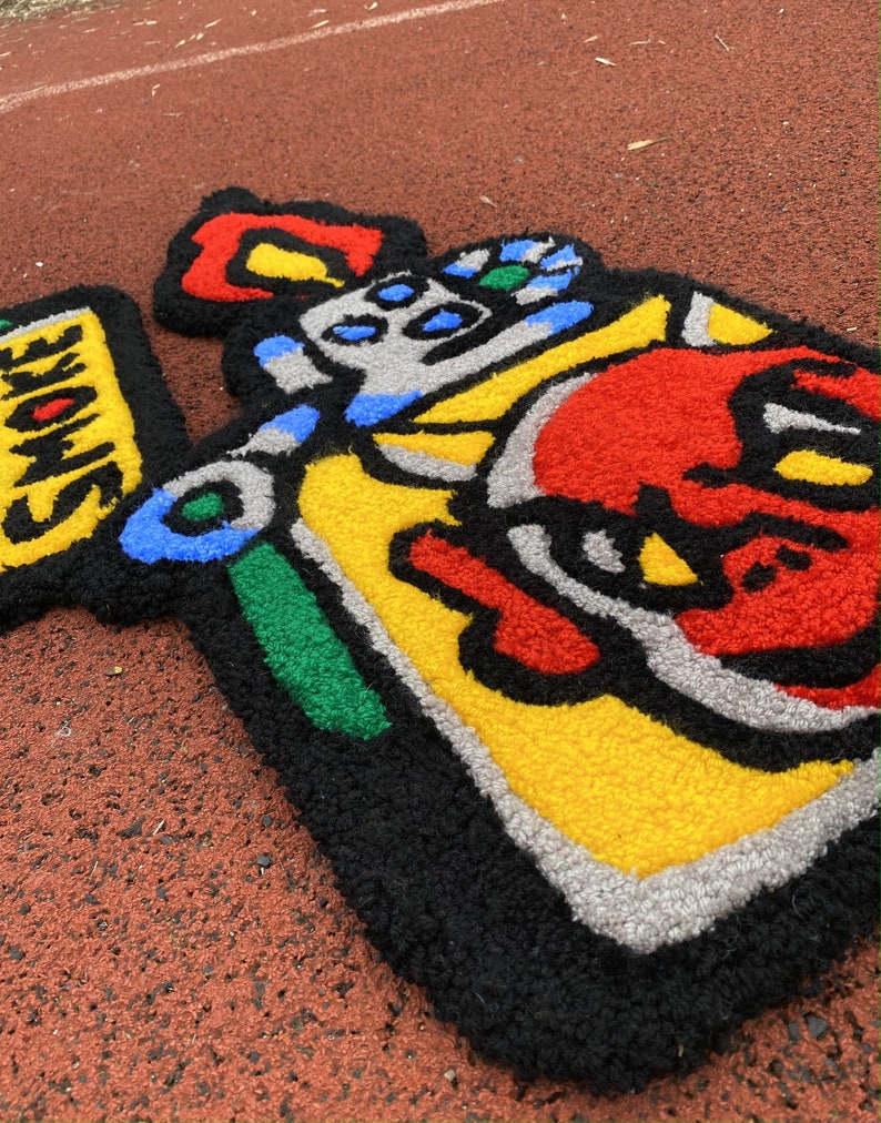 K&ouml;nnte beinhalten: Ein farbenfroher, handgefertigter Teppich mit einem Cartoon-Design. Der Teppich hat einen schwarzen Umriss mit roten, gelben, blauen, gr&uuml;nen und grauen Abschnitten. Das Wort "SMOKE" ist auf gelbem Hintergrund zu sehen. Der Teppich liegt auf einer roten Oberfl&auml;che.
