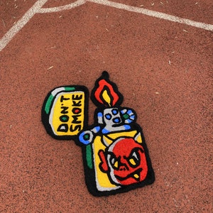 K&ouml;nnte beinhalten: Ein farbenfroher Teppich in Form eines Feuerzeugs mit einem Teufelsgesicht-Design. Der Teppich hat einen gelben K&ouml;rper, ein rotes Teufelsgesicht und eine Flamme. Der Text "DON'T SMOKE" ist auf dem Teppich zu sehen.