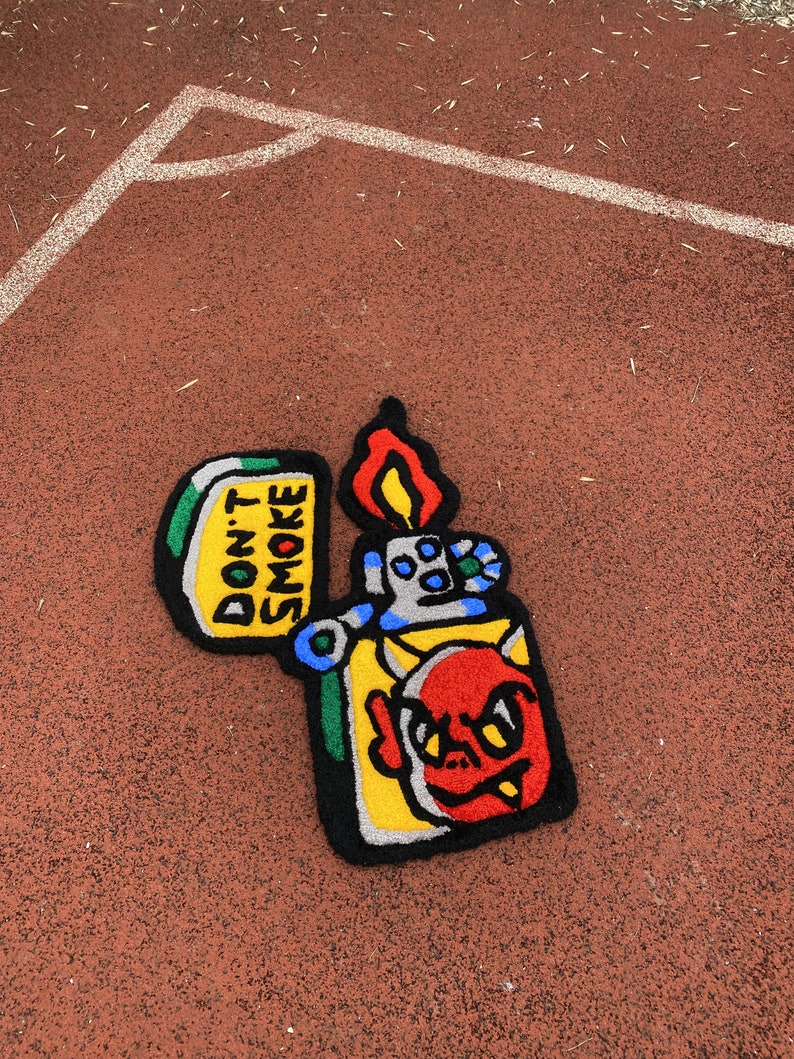 K&ouml;nnte beinhalten: Ein farbenfroher Teppich in Form eines Feuerzeugs mit einem Teufelsgesicht-Design. Der Teppich hat einen gelben K&ouml;rper, ein rotes Teufelsgesicht und eine Flamme. Der Text "DON'T SMOKE" ist auf dem Teppich zu sehen.