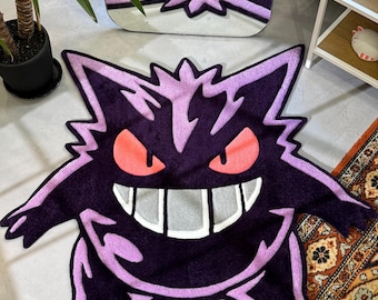 Puede incluir: Una alfombra morada y rosa con una imagen de dibujos animados de un fantasma con una amplia sonrisa y ojos grandes. La alfombra está hecha de un material suave y mullido.