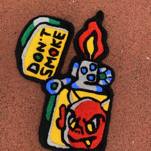 K&ouml;nnte beinhalten: Ein farbenfroher Teppich in Form eines Feuerzeugs mit einem Cartoon-Teufel und dem Text "DON'T SMOKE". Der Teppich hat ein Farbschema aus Gelb, Rot, Blau, Gr&uuml;n und Schwarz. Die Flamme ist rot und gelb.