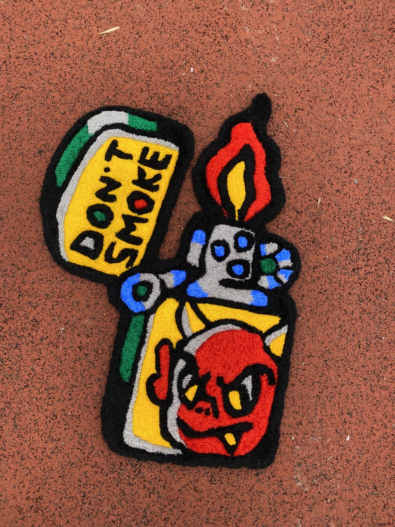 K&ouml;nnte beinhalten: Ein farbenfroher Teppich in Form eines Feuerzeugs mit einem Cartoon-Teufel und dem Text "DON'T SMOKE". Der Teppich hat ein Farbschema aus Gelb, Rot, Blau, Gr&uuml;n und Schwarz. Die Flamme ist rot und gelb.