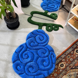May include: A vibrant blue rug shaped like a stylized fruit, with a green stem. The rug has a textured, plush surface with swirling patterns. The rug is on a light-colored floor, with a patterned rug in the corner.