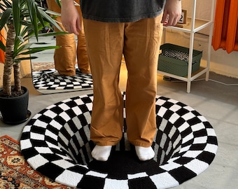 May include: A round rug featuring a black and white checkered pattern that creates a three-dimensional optical illusion of a deep hole. The rug is positioned on a floor, with a pair of feet in white socks standing directly in the center of the design.