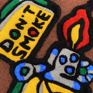 K&ouml;nnte beinhalten: Ein farbenfroher Teppich in Form eines Feuerzeugs mit der Aufschrift "DON'T SMOKE". Der Teppich hat einen gelben K&ouml;rper, eine rote und gelbe Flamme sowie blaue und graue Details. Das Design ist schwarz umrandet.
