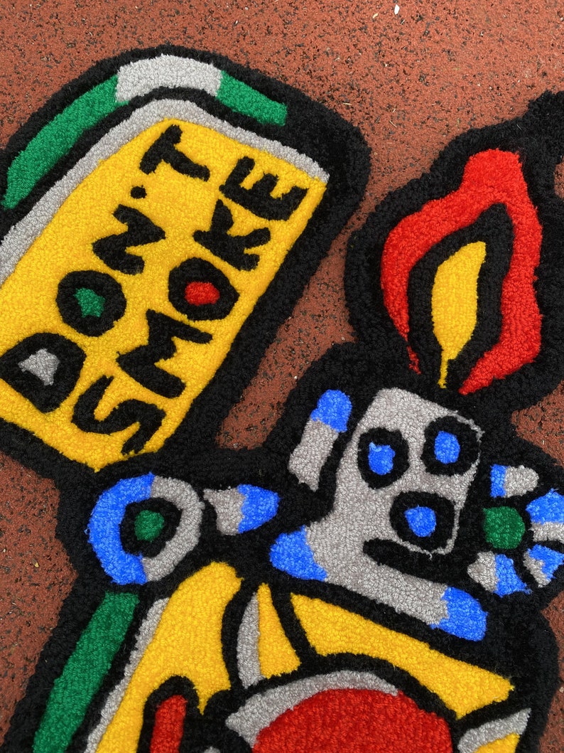 K&ouml;nnte beinhalten: Ein farbenfroher Teppich in Form eines Feuerzeugs mit der Aufschrift "DON'T SMOKE". Der Teppich hat einen gelben K&ouml;rper, eine rote und gelbe Flamme sowie blaue und graue Details. Das Design ist schwarz umrandet.