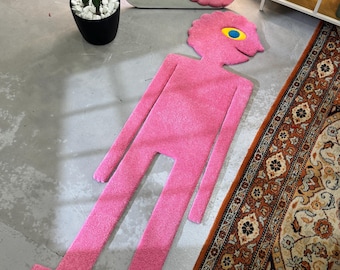 May include: A pink, human-shaped rug with a textured surface. The rug features a stylized head with a single large eye, which has a yellow iris and a blue pupil. The body is elongated with long arms and legs, creating a distinct, flat, anthropomorphic silhouette.