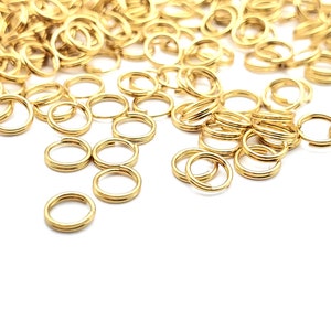 May include: A large pile of gold jump rings, with a few smaller rings scattered on a white surface.