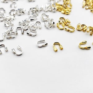 May include: A close-up of a collection of gold and silver crimp beads. The beads are arranged in two groups, with the gold beads on the right and the silver beads on the left. The beads are small and round, with a hole in the center. They are used to secure the ends of jewelry.
